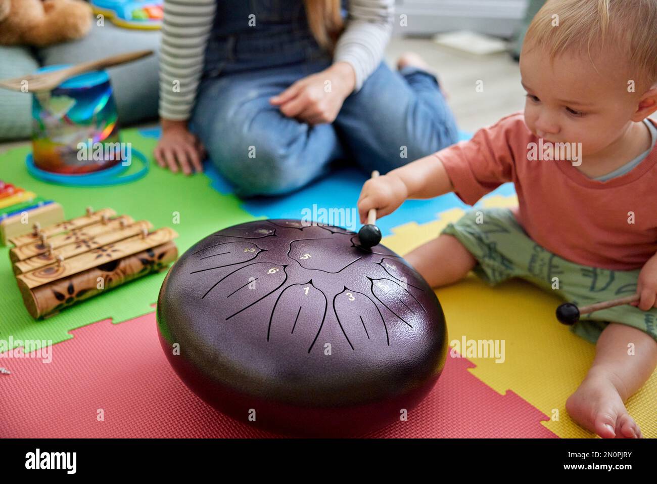 Toddler playing with musical instrument in playroom with mother sitting nearby Stock Photo