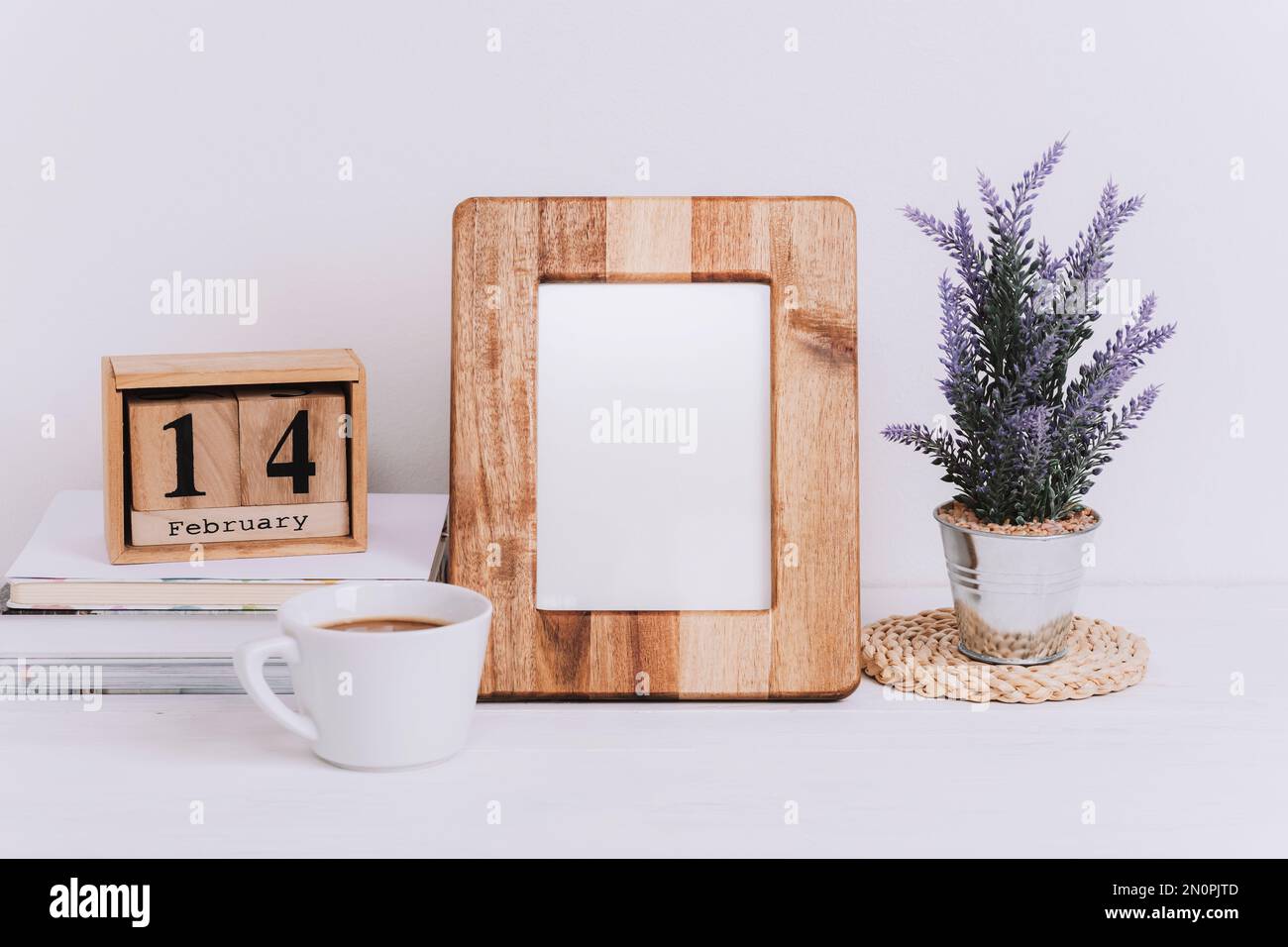 Mockup wooden picture frame and block calendar with Valentines day holiday date. Coffee cup, flowers in pot on white table. Copy space. Stock Photo