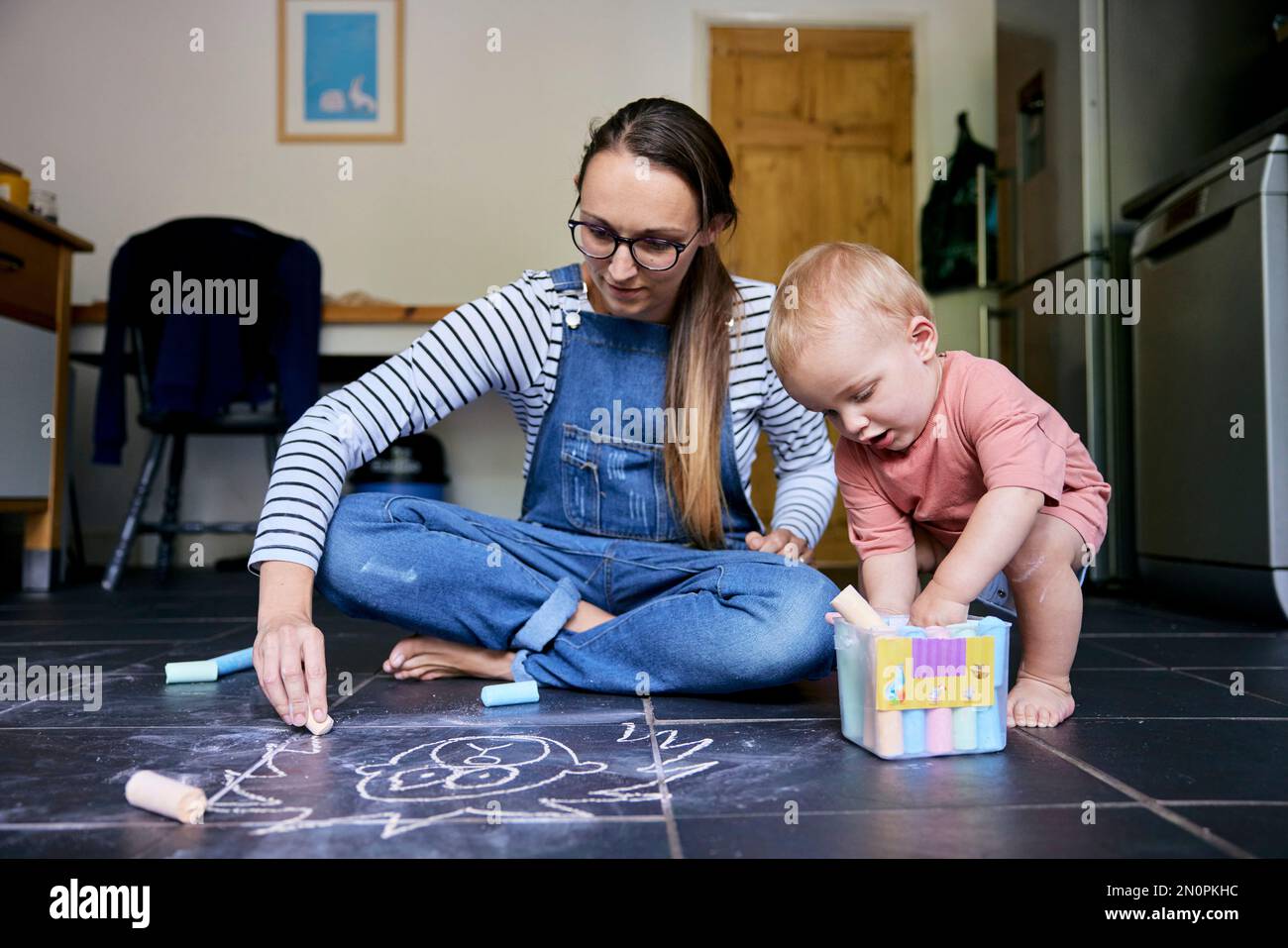 Mother and toddler drawing with chalks together on a tiled kitchen floor Stock Photo