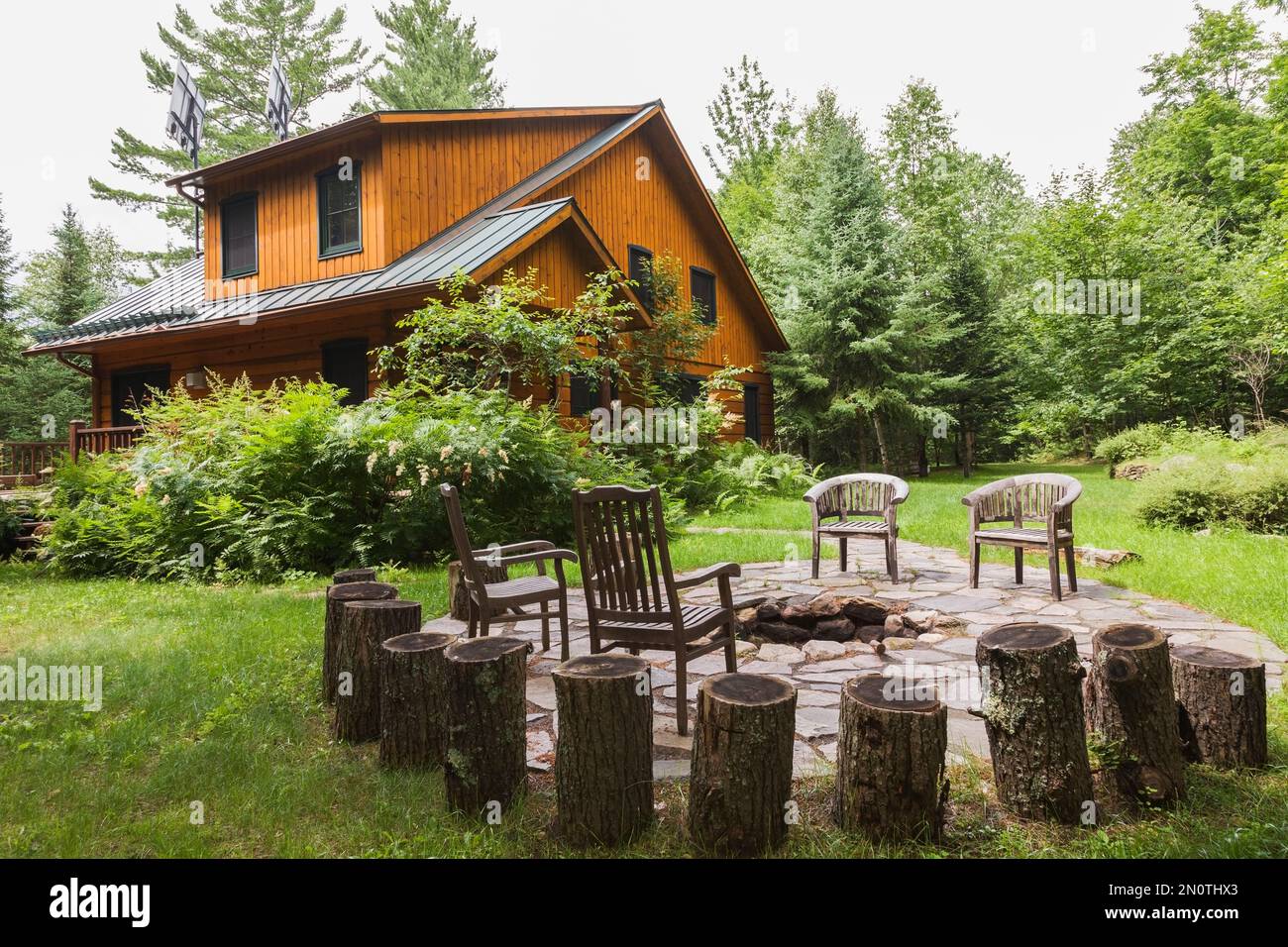 Milled cottage style flat log profile home facade with porch and landscaping plus firepit surrounded by flagstones in summer. Stock Photo