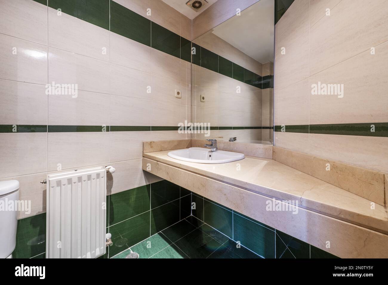 Bathroom with chunky cream marble countertop with white porcelain inset sink under frameless mirror and green tiled flooring Stock Photo