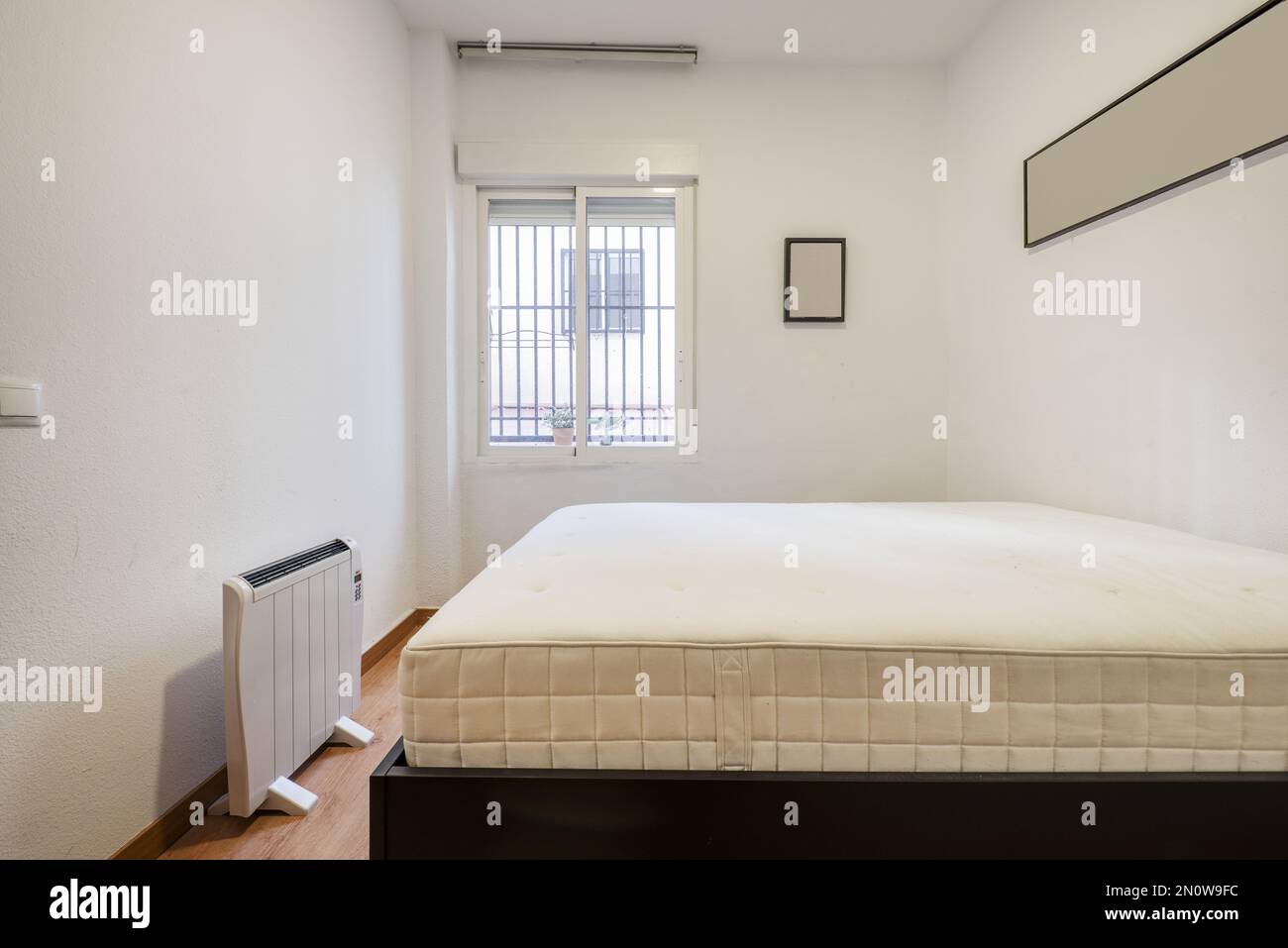 Bedroom mattress without bedding, an electric radiator and window with metal bars Stock Photo