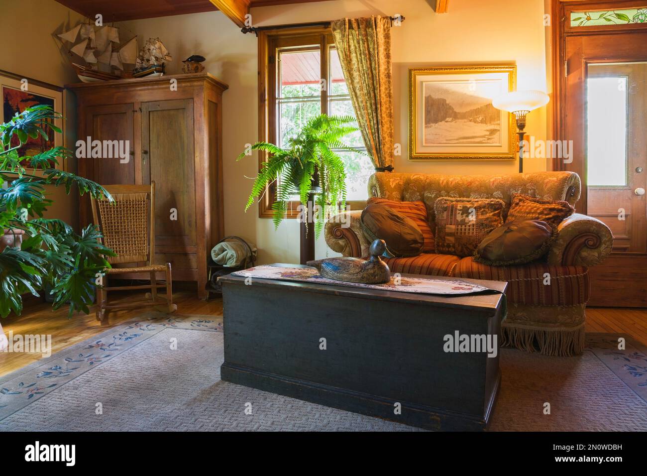 Family room with antique wooden storage chest coffee table, upholstered sofa, woven seat rocking chair and pinewood armoire inside old circa 1850 home. Stock Photo