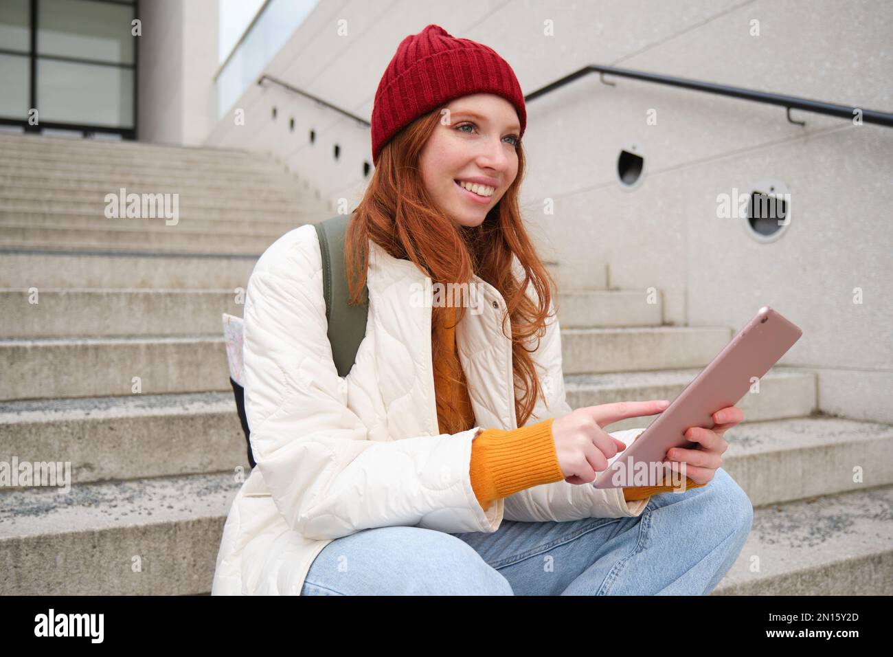 Happy stylish redhead girl, student in red hat, holds digital tablet, uses social media app, searches something online, connects to wifi. Stock Photo