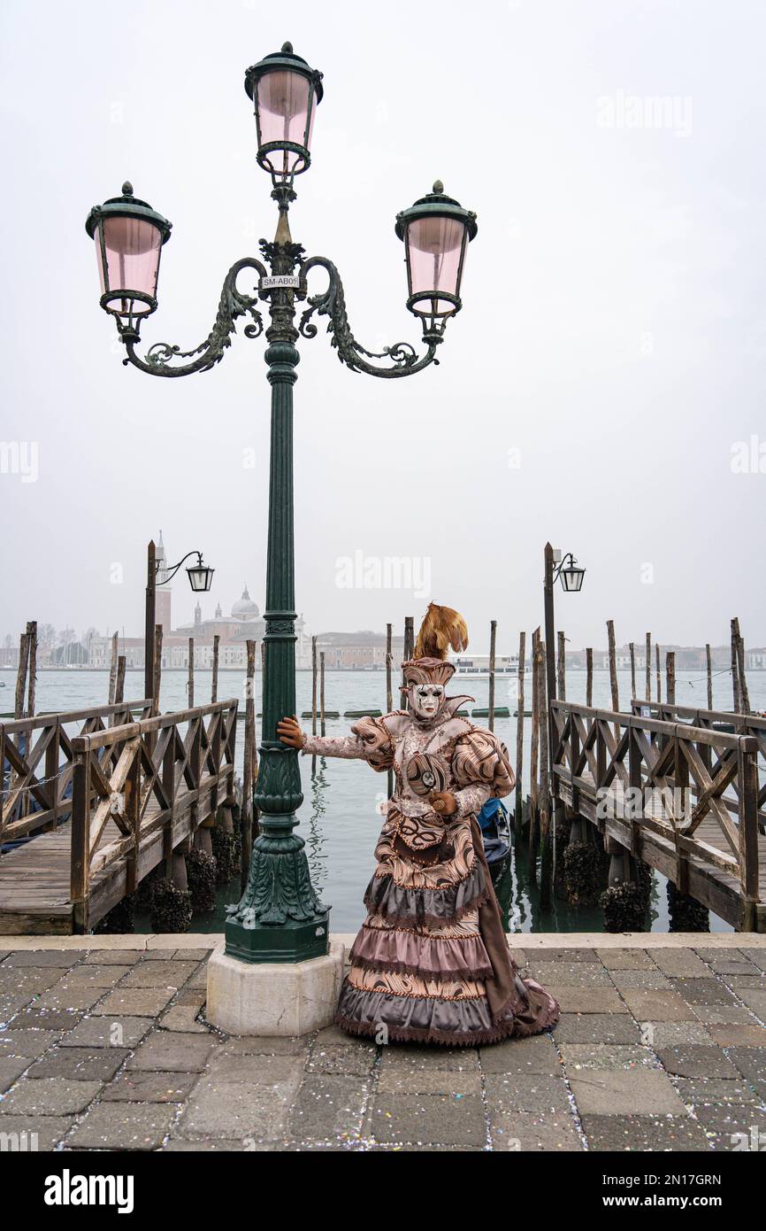 A woman in a carnival costume, a mask, a hat with feathers, a mirror in her hand, stands leaning on a lamppost against the background of the pier Stock Photo