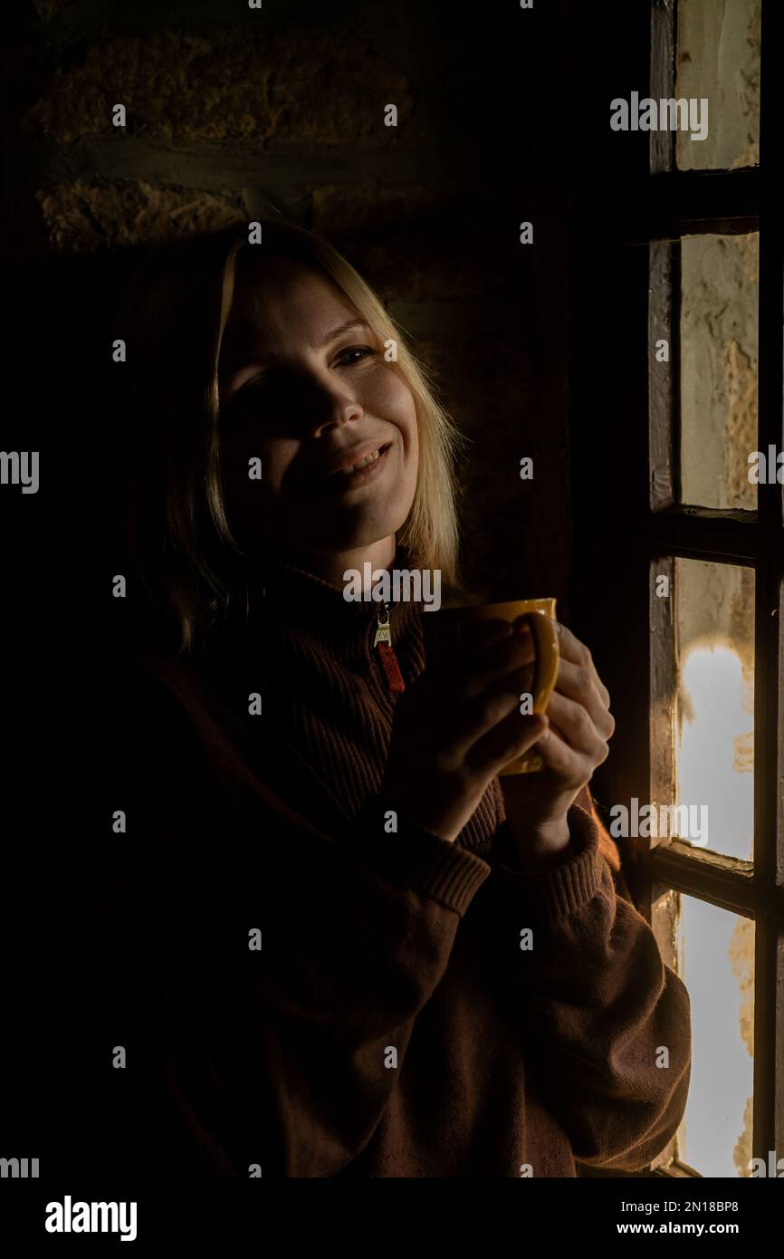 A woman smiles and drinks coffee by the window in the dark. A woman in ...