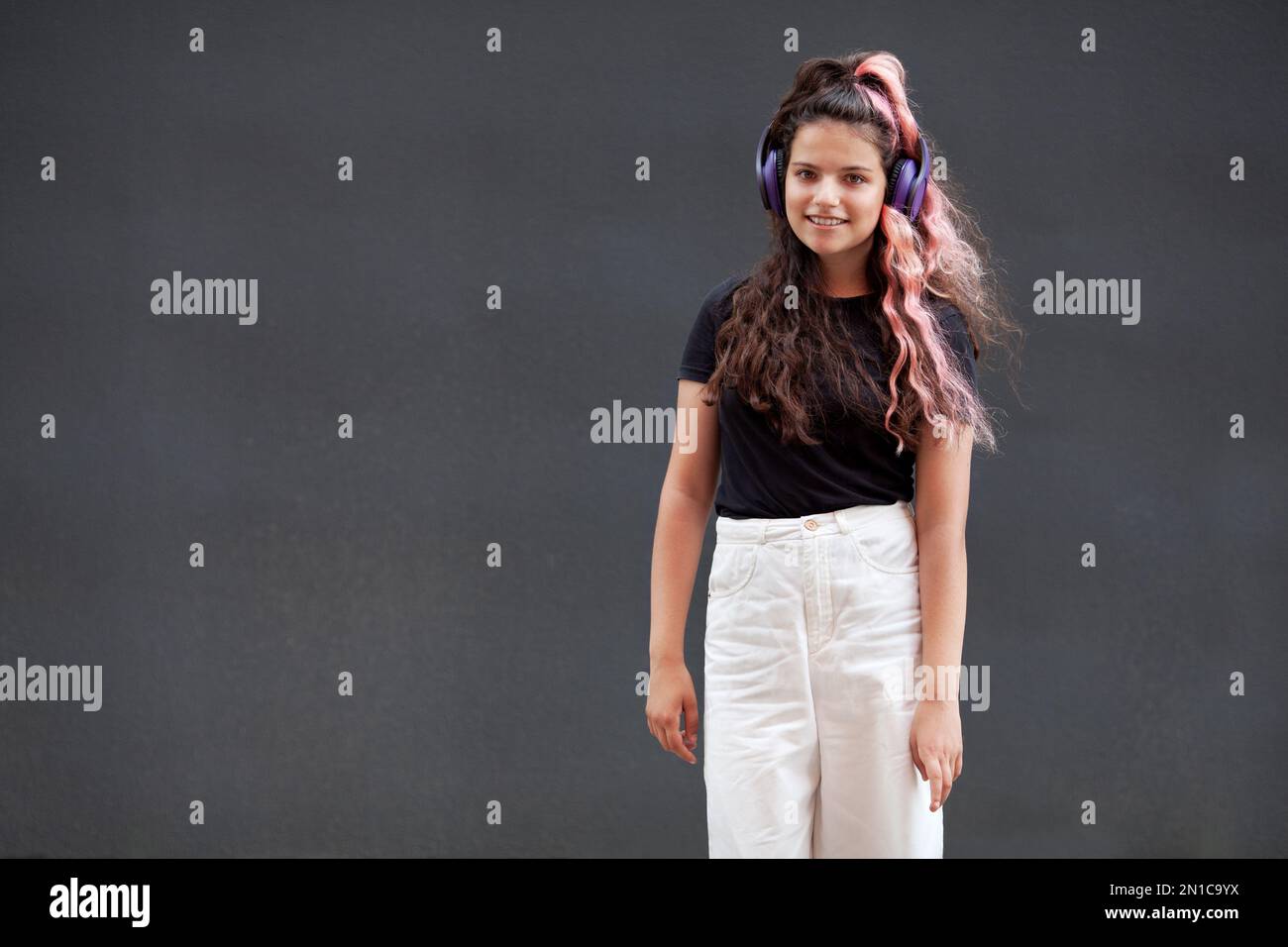 Young happy Girl with colored long pink hair listen to music with headphones, smiling and looking at camera isolated on dark gray background Stock Photo