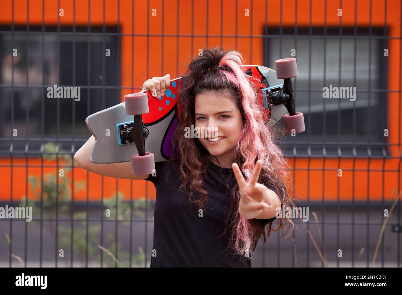 Colorful portrait happy young girl with skateboard smiling, looking at camera and showing peace or victory gesture Stock Photo