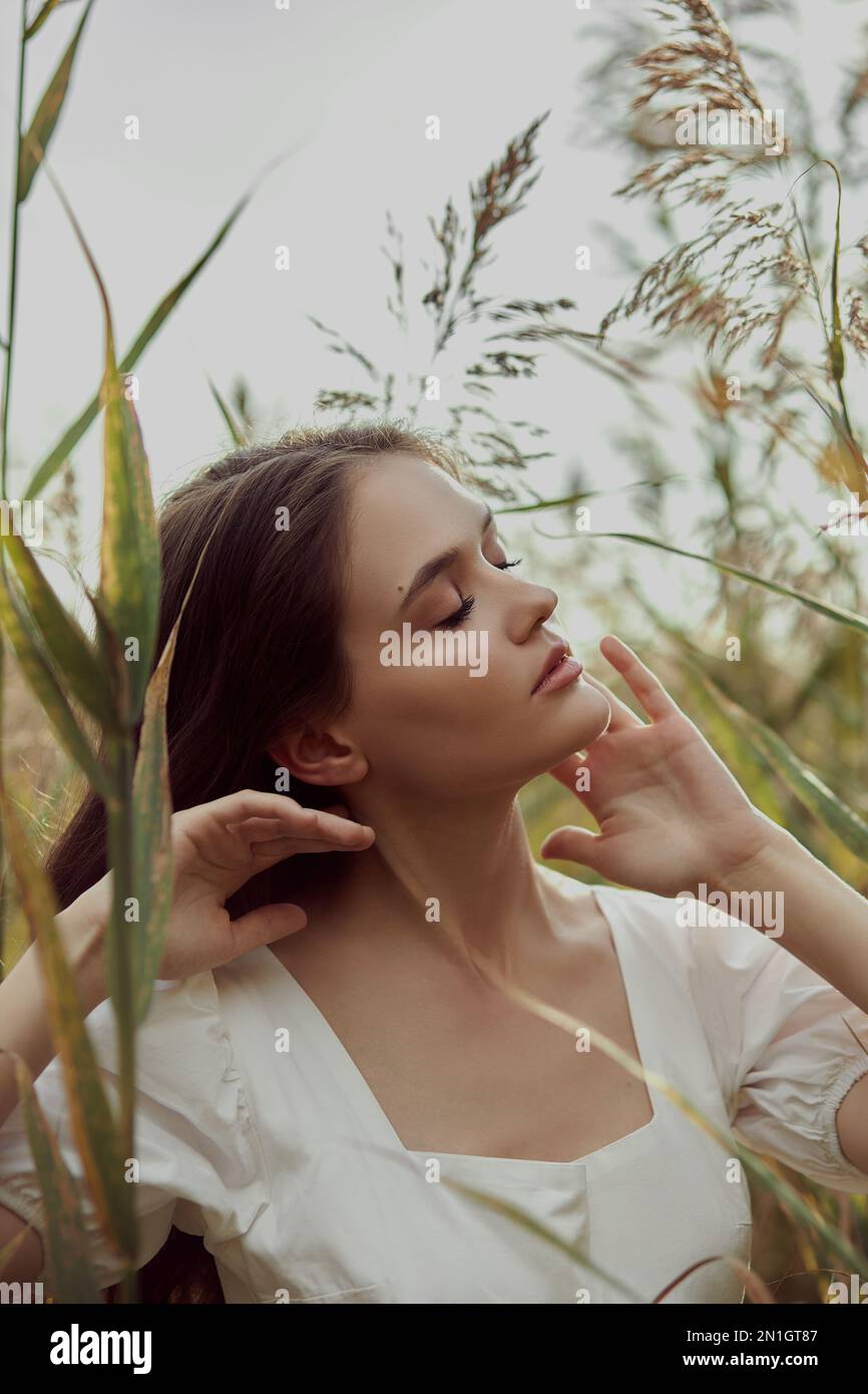 Beautiful young woman in white summer dress is sitting in tall grass in rural field. Portrait of romantic girl at sunset, warm sun, natural beauty wom Stock Photo