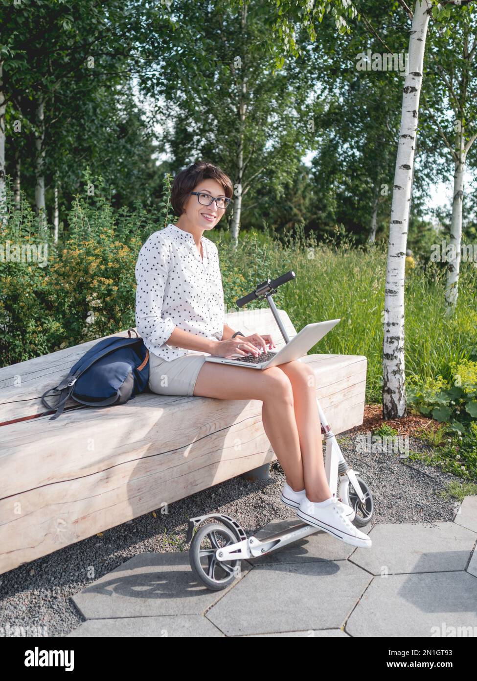 Smiling woman works with laptop on wooden bench in urban park. Summer vibes. Modern workplace for freelancers. Student educates online from outdoors. Stock Photo