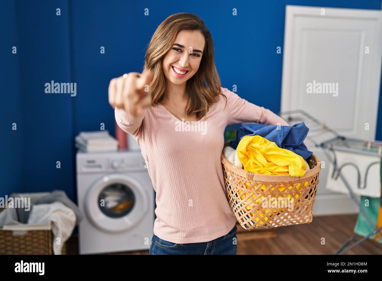 Young woman holding laundry basket pointing to you and the camera with ...
