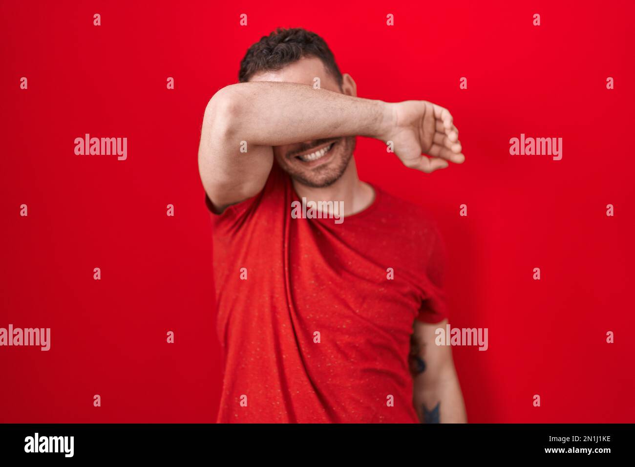 Young hispanic man standing over red background covering eyes with arm ...