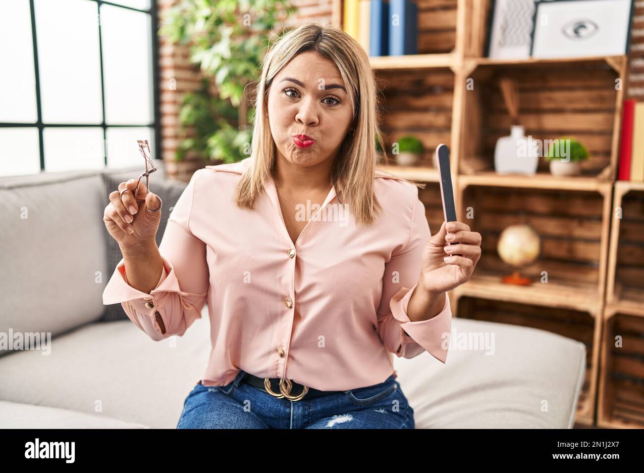 Young hispanic woman holding eye lashes curler and nail file puffing cheeks with funny face. mouth inflated with air, catching air. Stock Photo