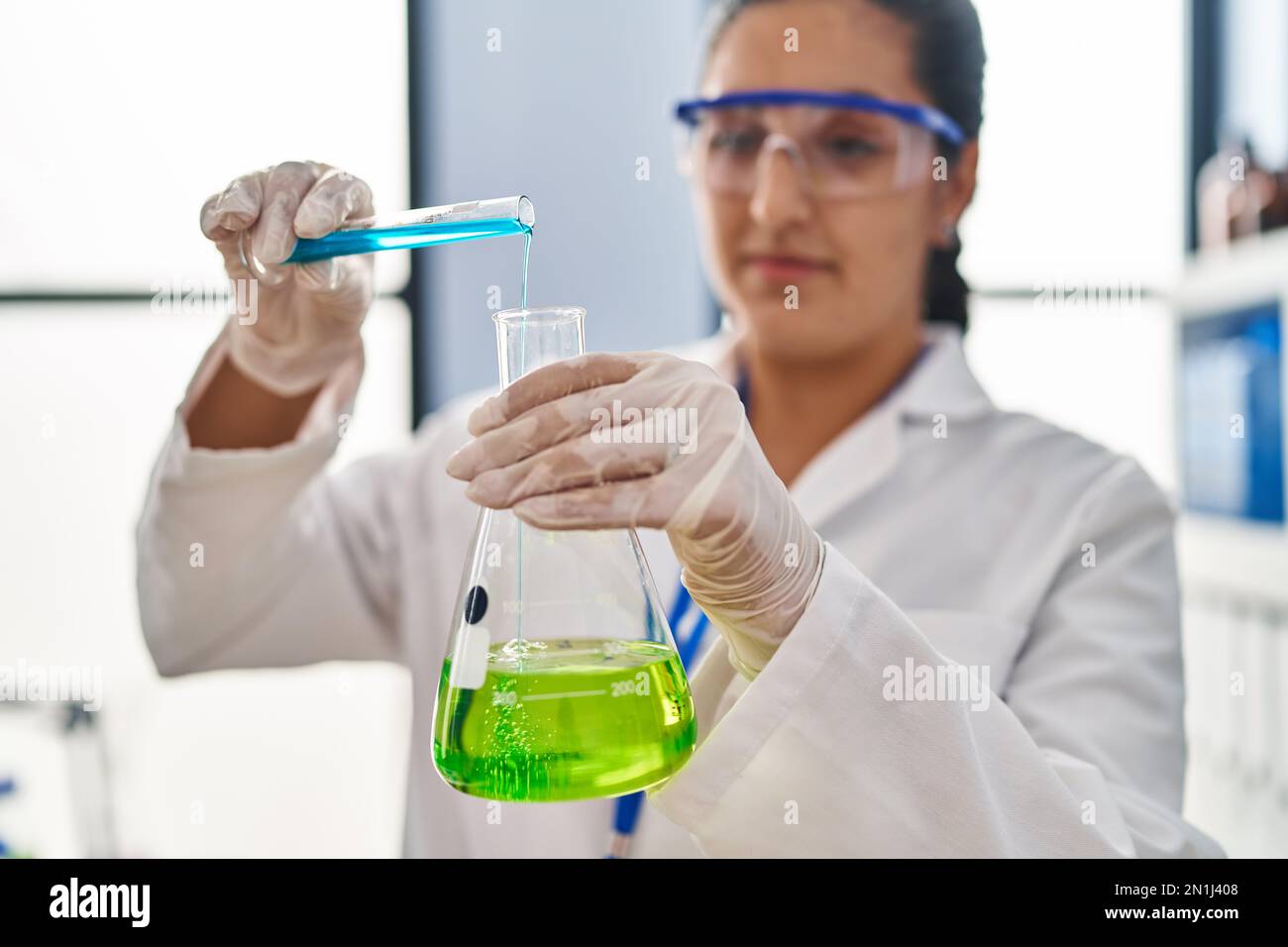 Young hispanic woman wearing scientist uniform pouring liquid on test
