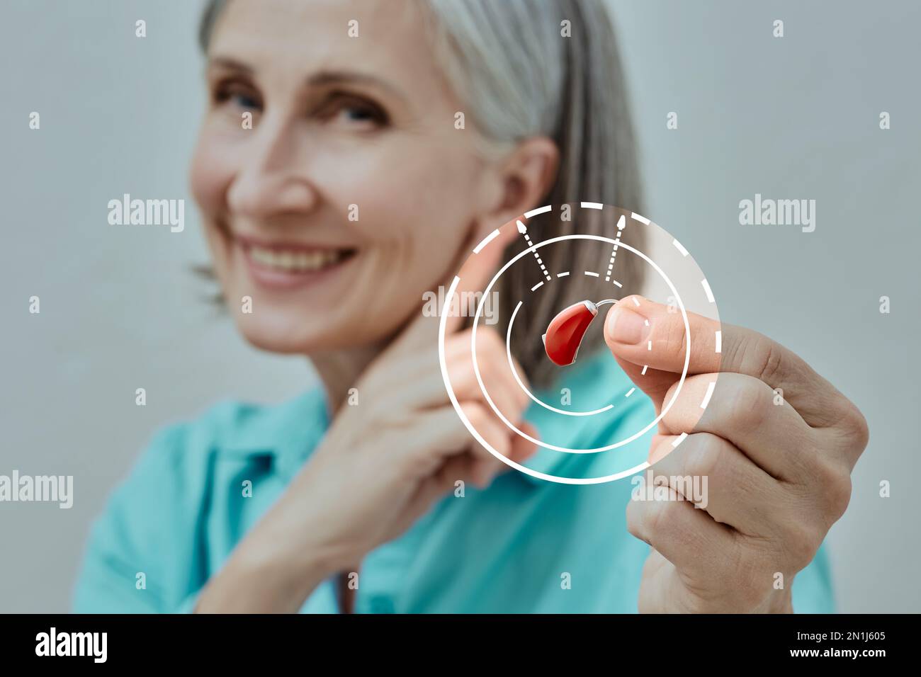 Mature woman holding BTE hearing aid in hand on foreground and points to her ear, concept. Hearing loss treatment Stock Photo