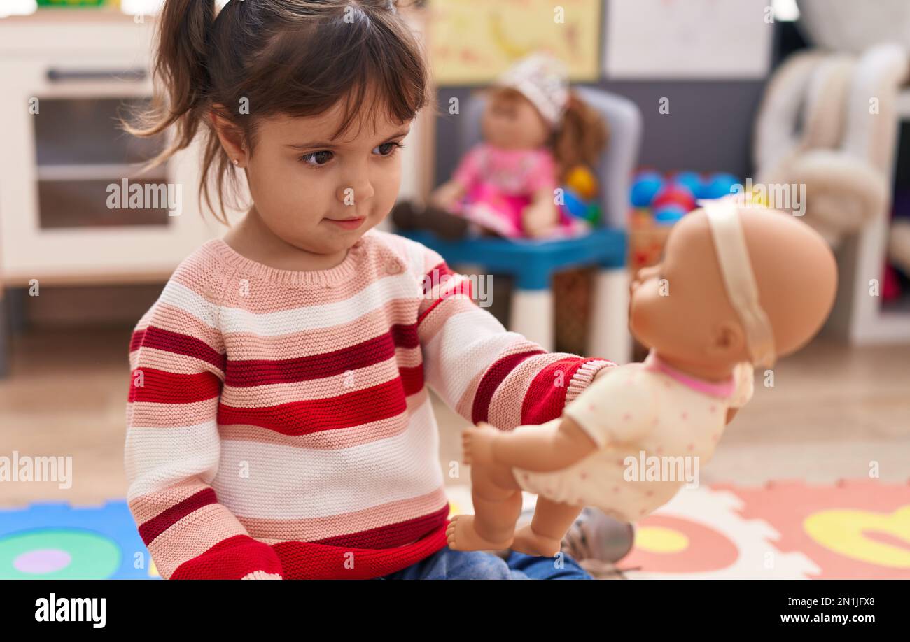 Adorable hispanic girl playing with baby doll standing at kindergarten ...