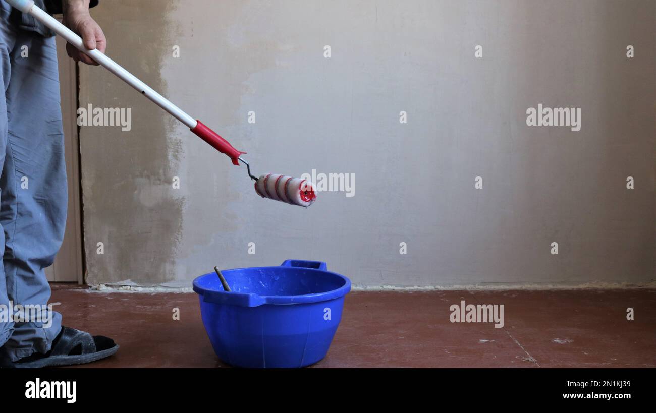worker with long handle roller and plastic bucket stands near plastered