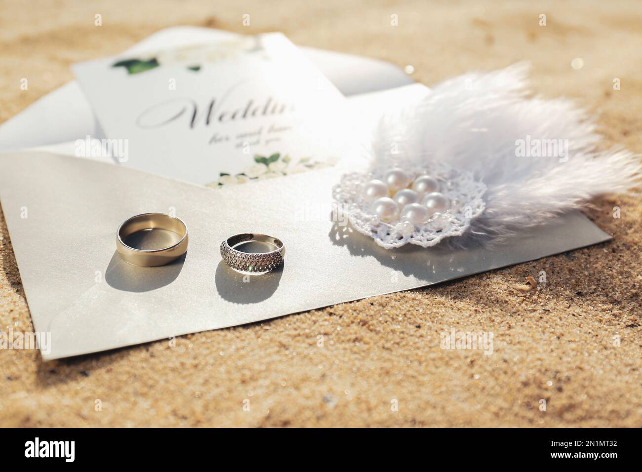 Envelope with wedding invitation, brooch and gold rings on sandy beach