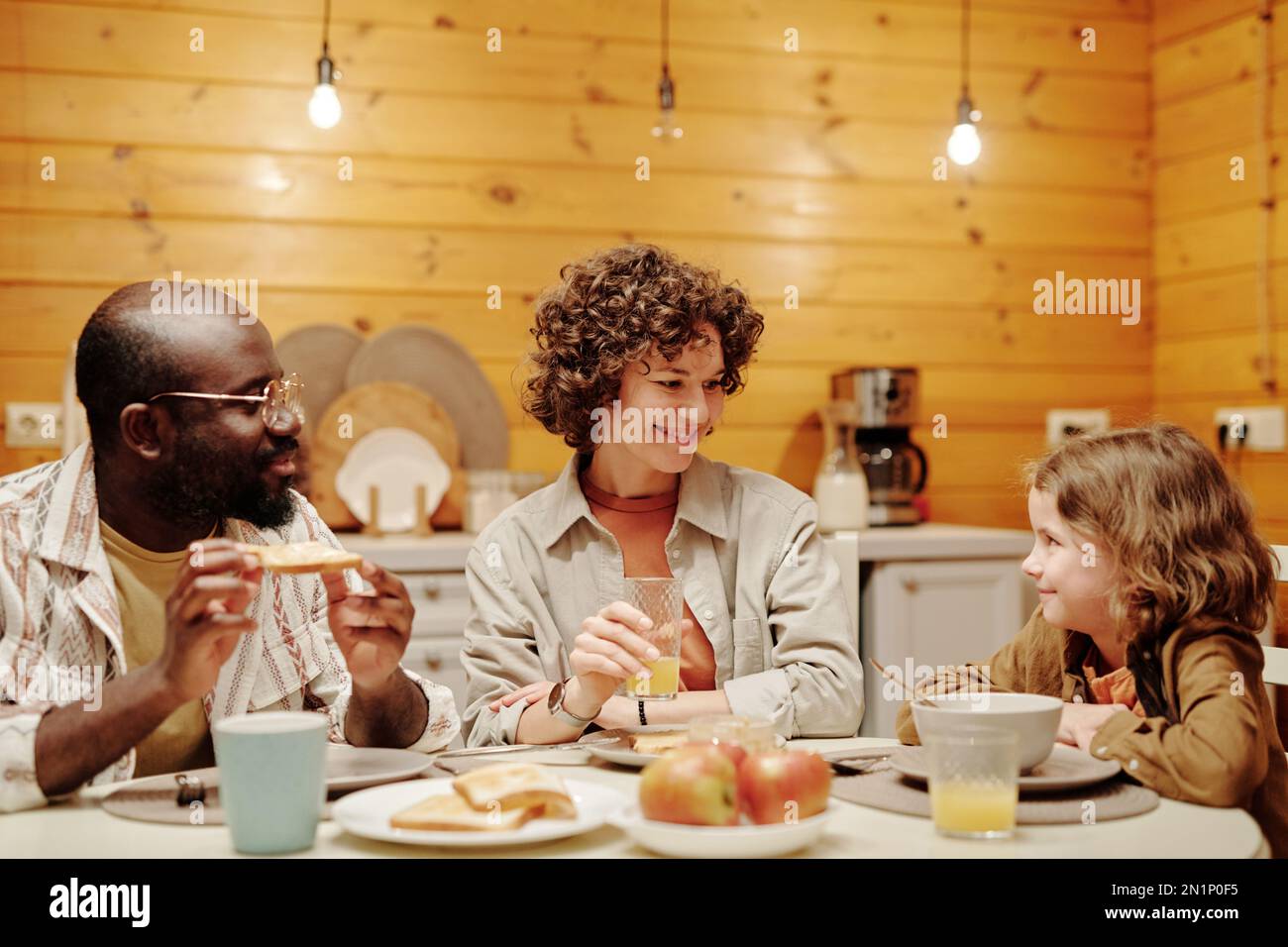 Young intercultural couple and their cute little son sitting by table served with sandwiches, apples and juice and having breakfast Stock Photo