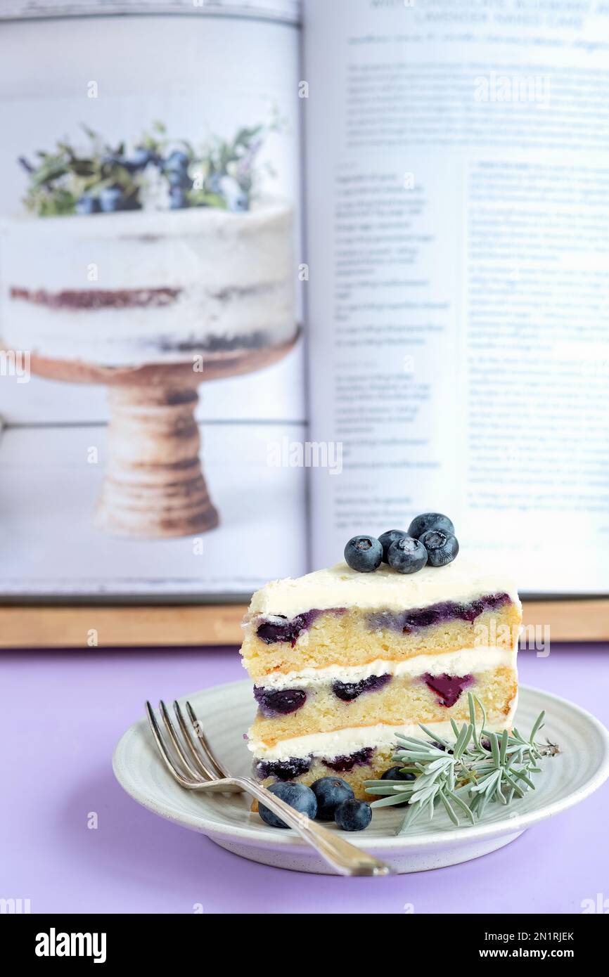 A slice of white chocolate, Blueberry Lavender cake, plated in front of the original cake recipe in the Canadian Maman cookbook Stock Photo