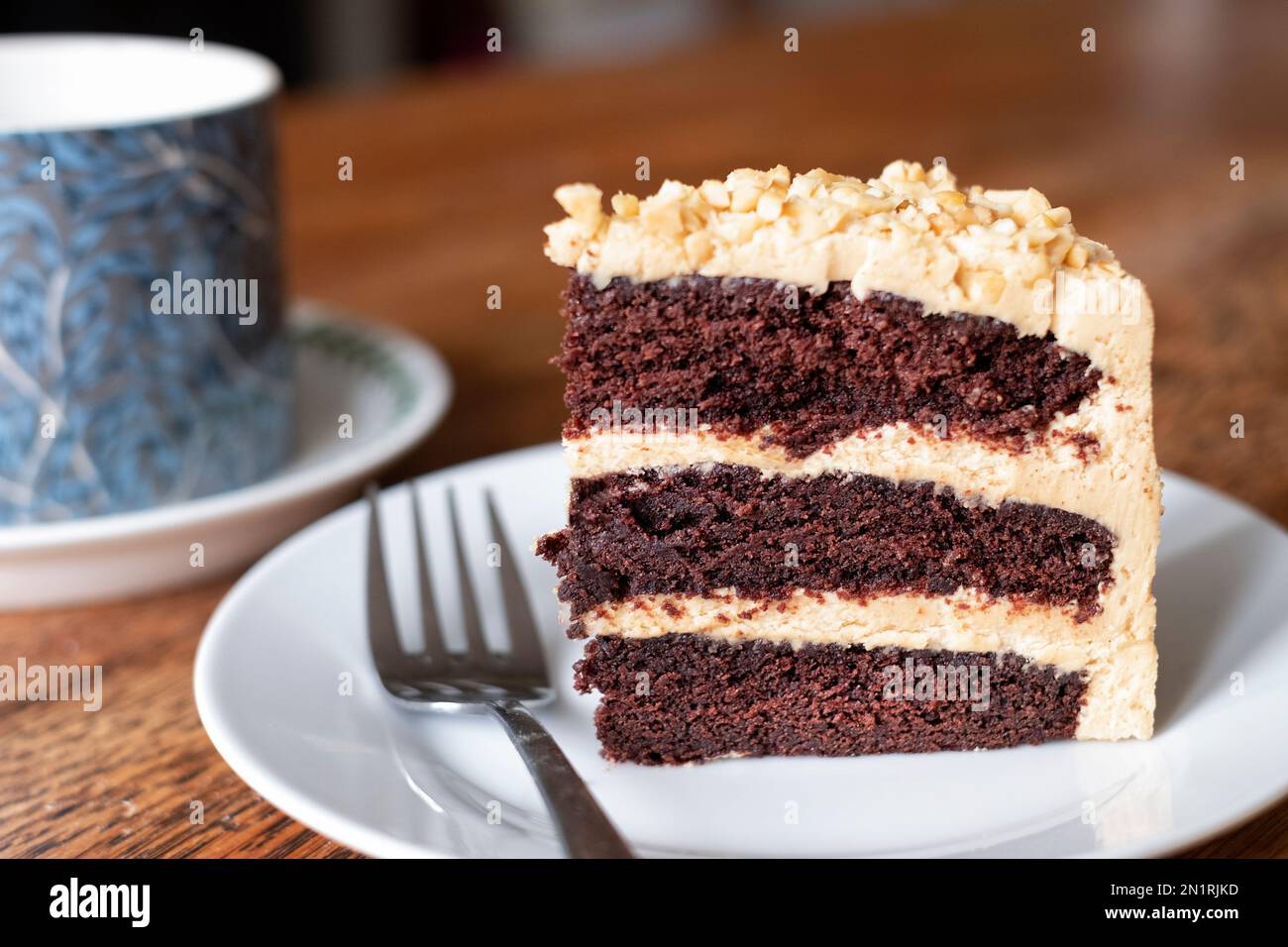 A slice of freshly baked, peanut butter and chocolate cake. The cake has a thick buttercream filling and is served on a side plate with a cup of tea Stock Photo