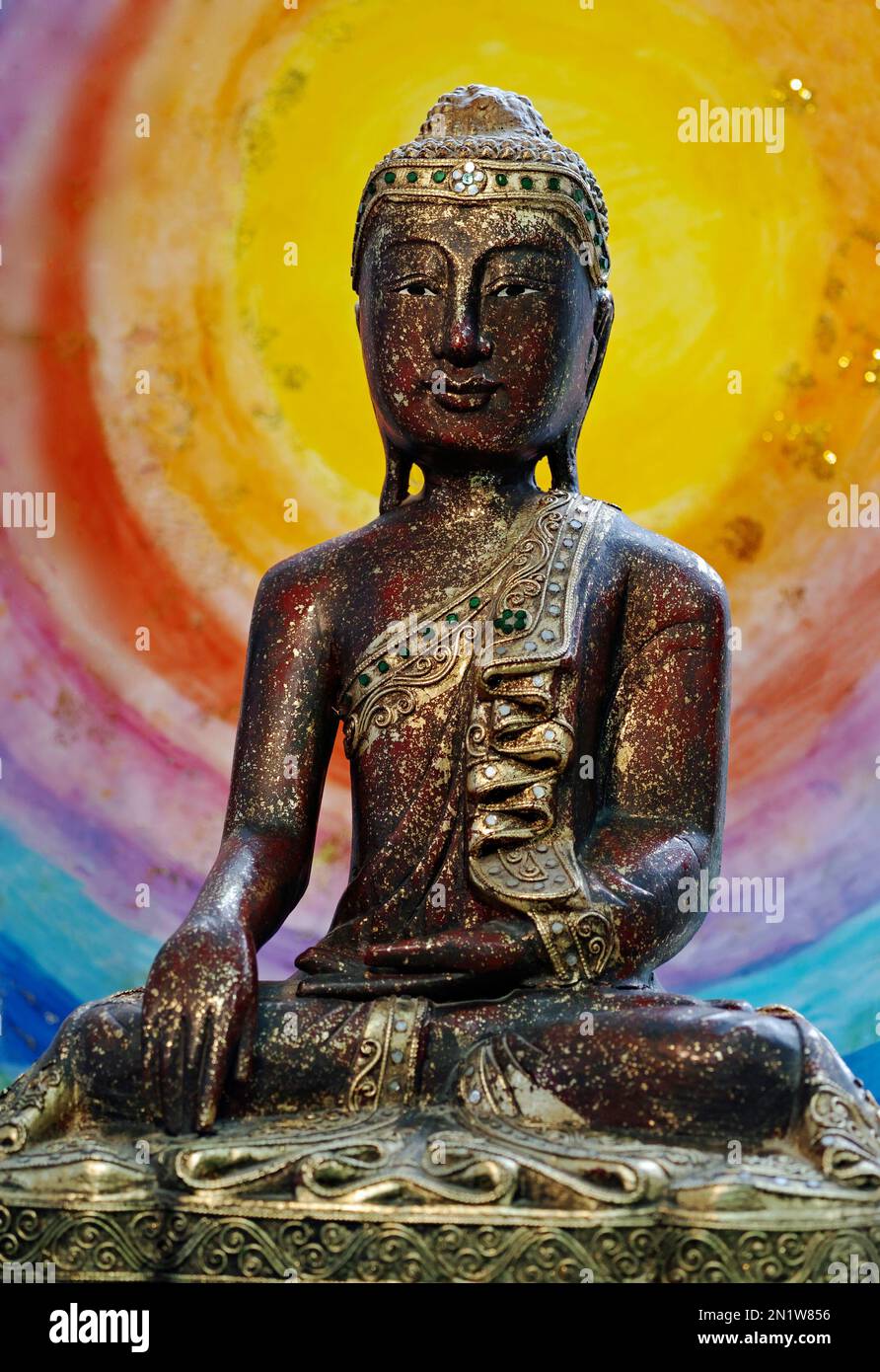 Buddha touching the earth, He is made of wood and decorated with gold. Probably made in India. In the background is a painting with rainbow colors. Stock Photo
