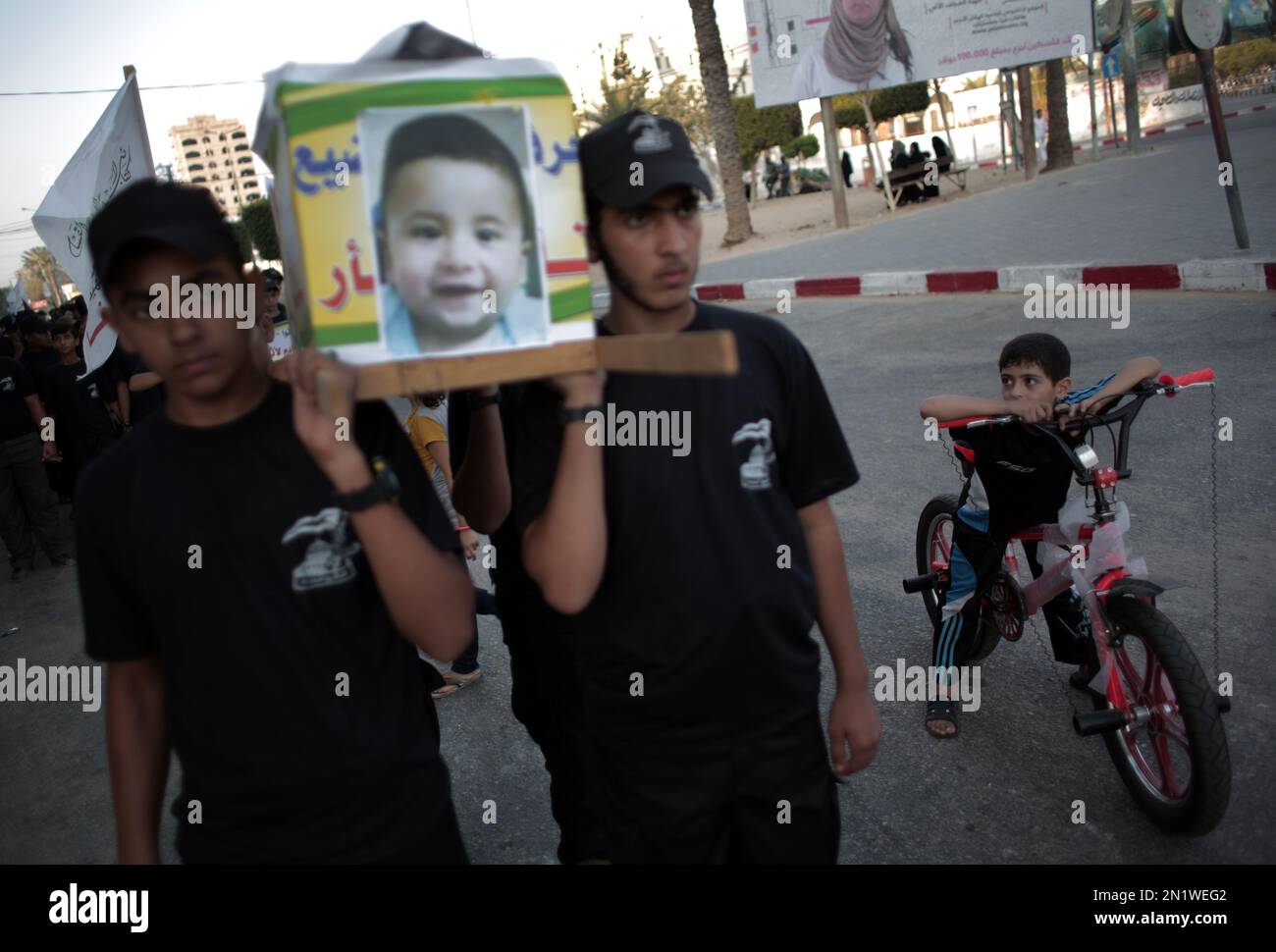 A Palestinian boy on his bicycle looks at youth carry a mock