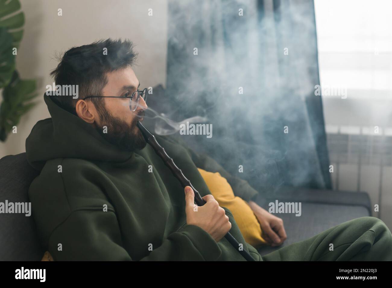 Bearded millennial or gen z man smoking hookah while relaxing on sofa
