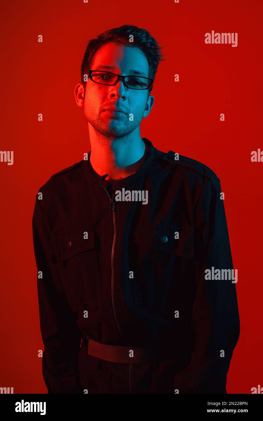 Studio Portrait of a handsome fashionable young caucasian brunette man wearing eyeglasses in a stylish jumpsuit at neon red light glowing background Stock Photo