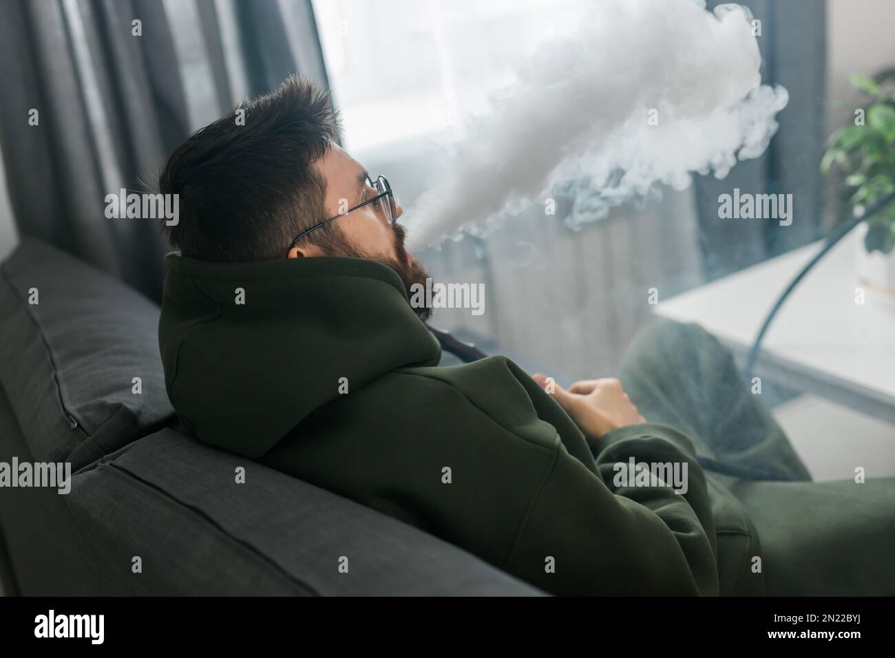 Bearded millennial or gen z man smoking hookah while relaxing on sofa