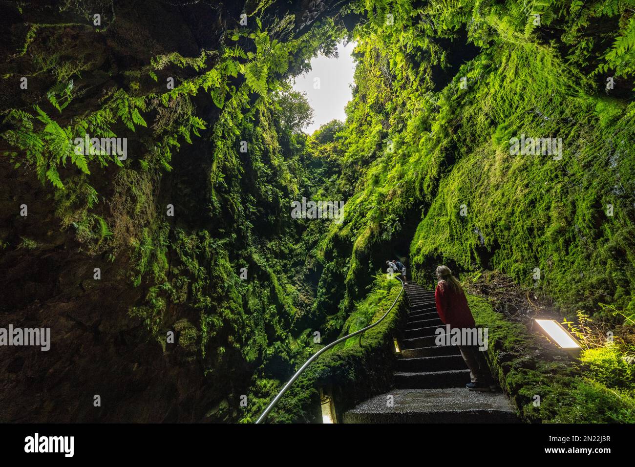 A tourist looks up inside the Algar do Carvão, a vertical lava tube dropping 300 feet from the surface accessible by a narrow staircase to a clear water pool at the bottom in the central mountains, Terceira Island, Azores, Portugal. Stock Photo