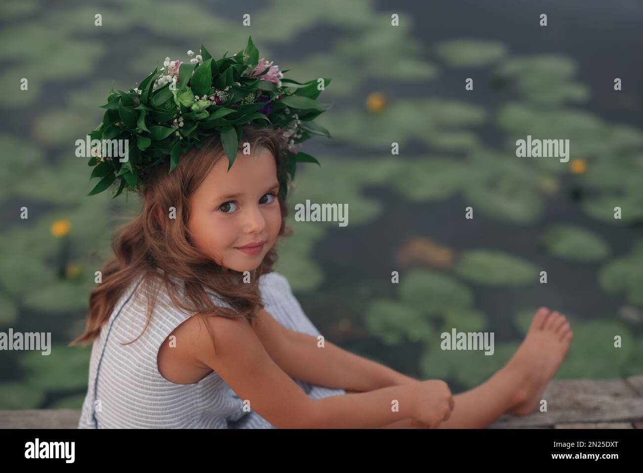 Cute little girl wearing wreath made of beautiful flowers near pond ...