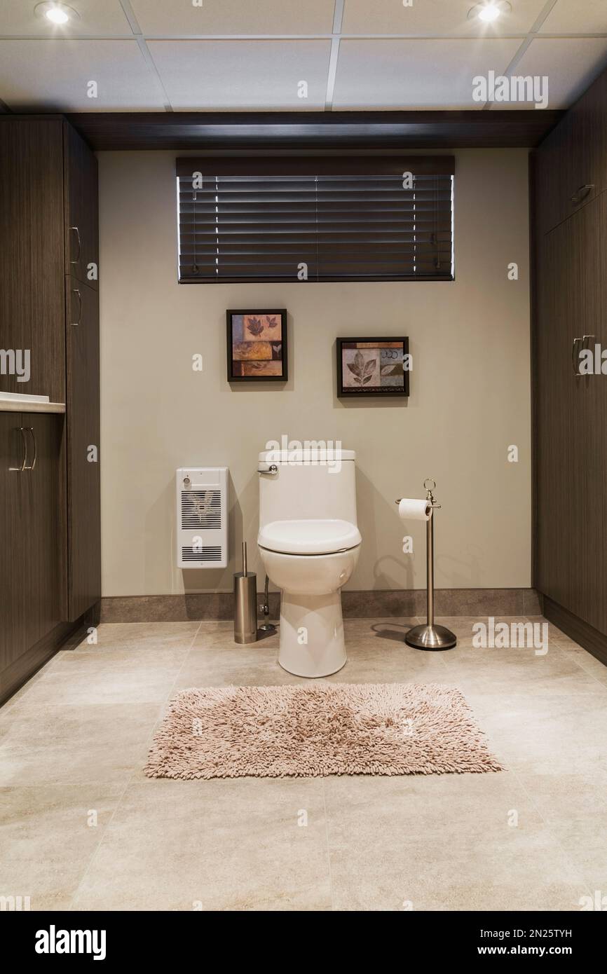 White porcelain toilet and brown wood veneer cabinets in laundry room bathroom in basement inside small home. Stock Photo