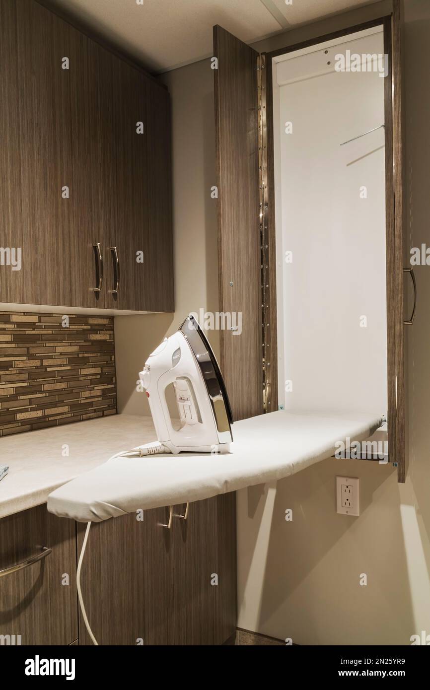 Clothes iron resting on top of fold out ironing board in laundry room bathroom with brown wood veneer cabinets in basement room inside small home. Stock Photo