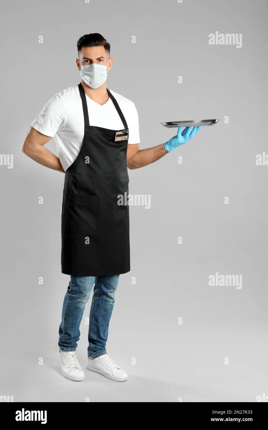Waiter in medical face mask with empty tray on light grey background ...