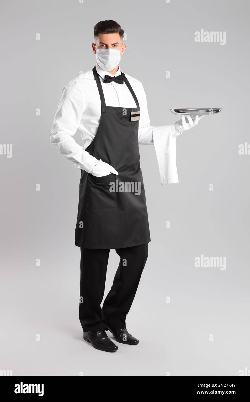 Waiter in medical face mask with empty tray on light grey background ...