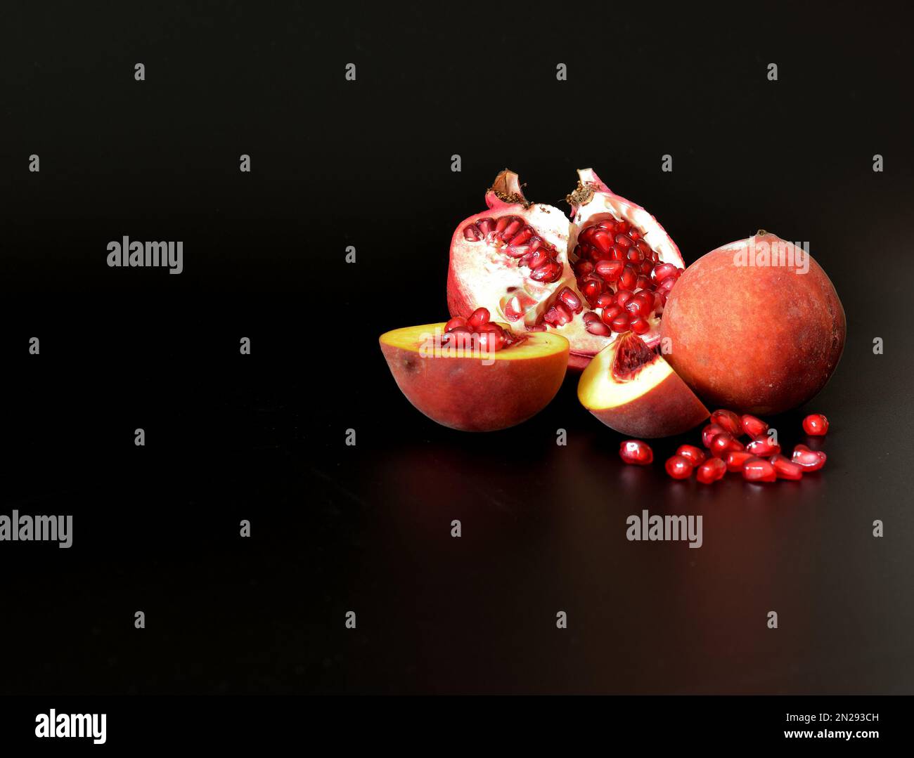 Broken pomegranate fruit with a scattering of seeds and pieces of a ripe peach on a black background. Close-up. Stock Photo