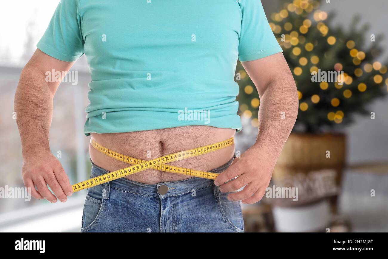 Overweight man measuring his waist in room decorated for Christmas after holidays, closeup Stock Photo