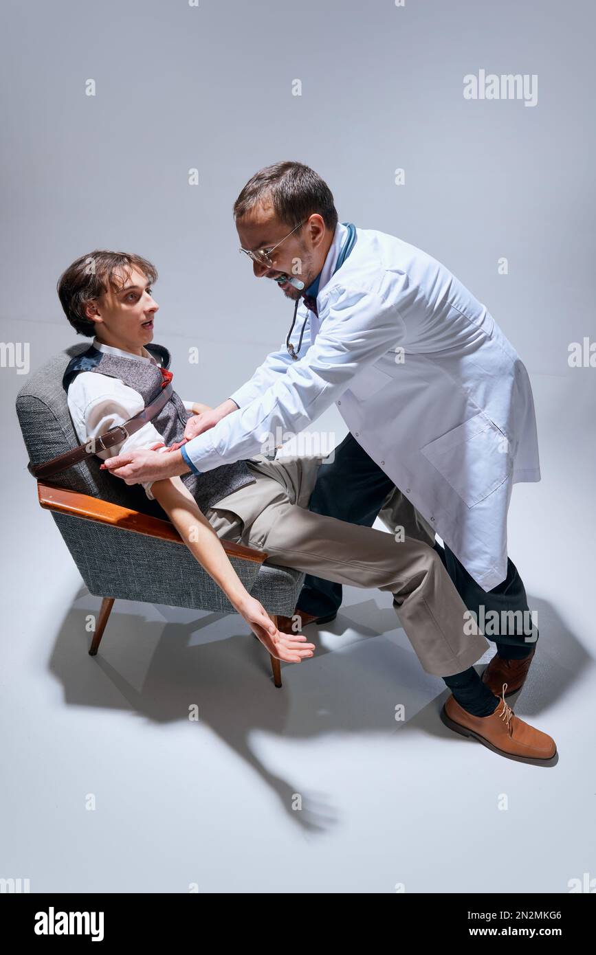 Crazy insane doctor, dentist tying young scared man on chair with belt  against grey studio background. Medicine and fear Stock Photo - Alamy