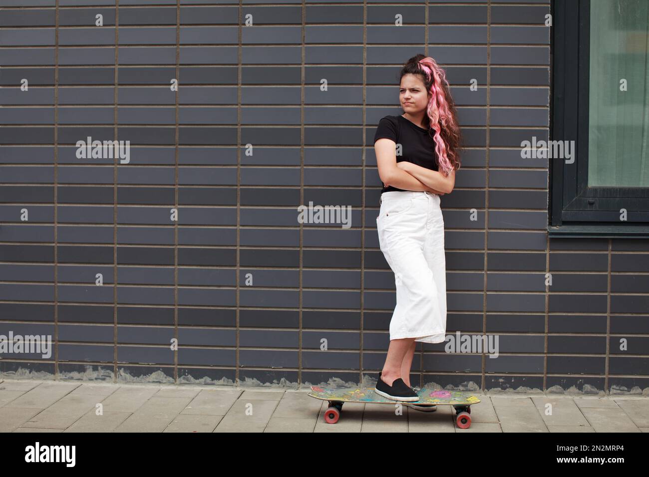 Sad teenage girl with skateboard stands full length on street next to wall of gray brick. Crisis of adolescence. Authentic lifestyle portrait of teena Stock Photo