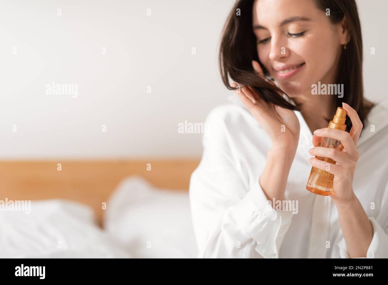 Glad caucasian millennial female sits on bed puts spray on hair, looks at ends, enjoy good morning, treatment Stock Photo