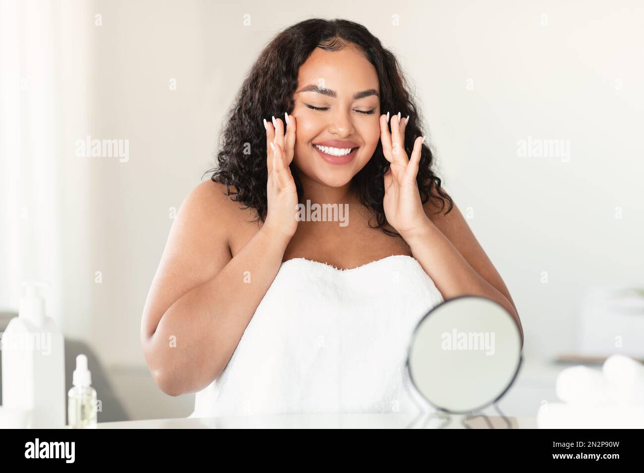 Facelift, beauty secrets concept. Happy plus size woman massaging her face, sitting in front of the mirror and smiling Stock Photo