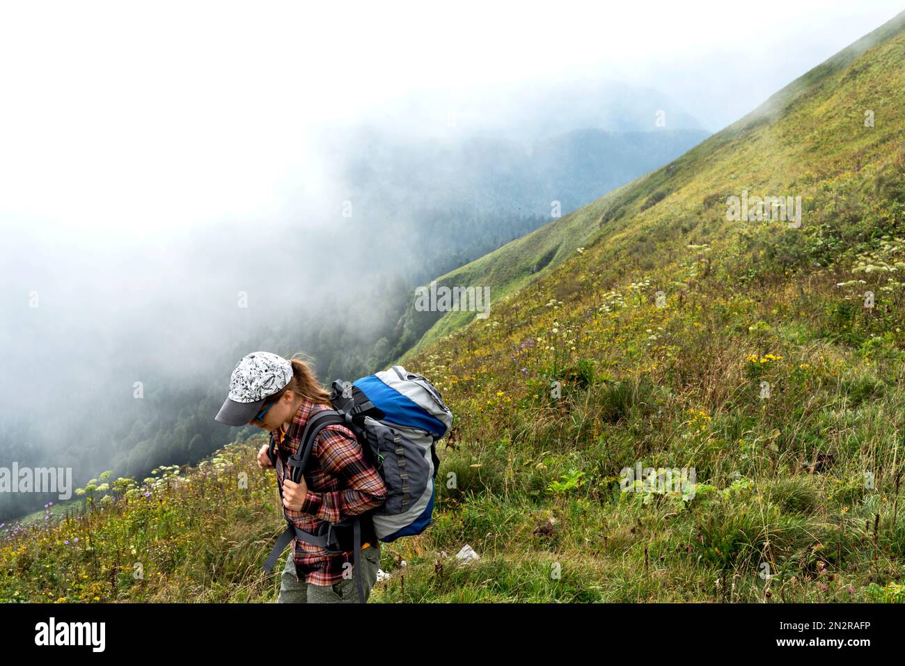 young woman hiker in plaid shirt and cap with big backpack walking in green mountains against clouds in summer healthy active lifestyle, outdoor activ Stock Photo