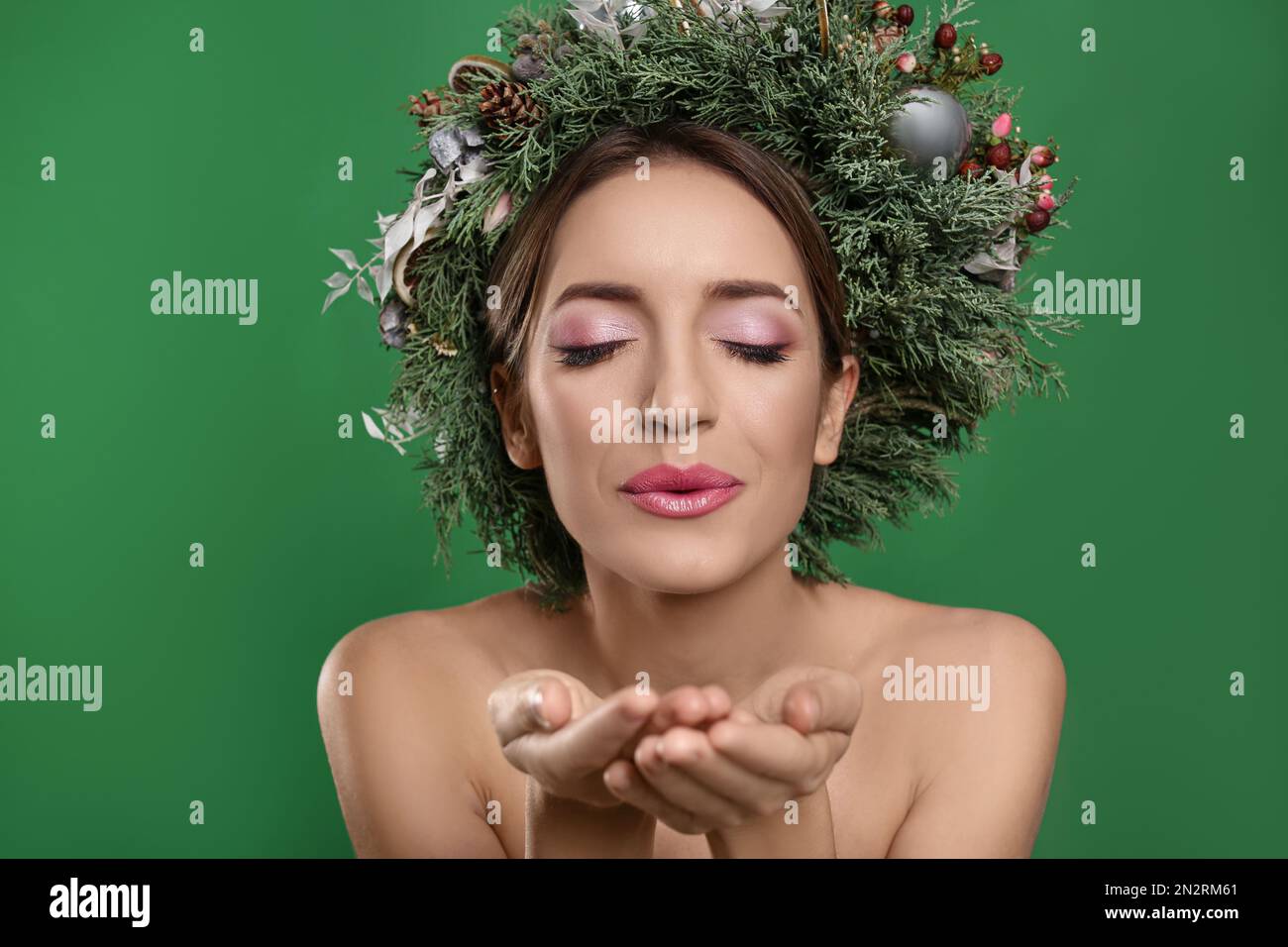 Beautiful young woman with Christmas wreath blowing kiss on green background Stock Photo