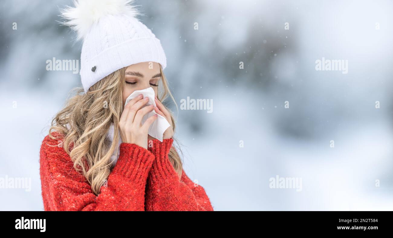Young woman with allergy symptom blowing nose. A sick girl in a warm knitted sweater and a woolen cap using a tissue in a winter park. Stock Photo