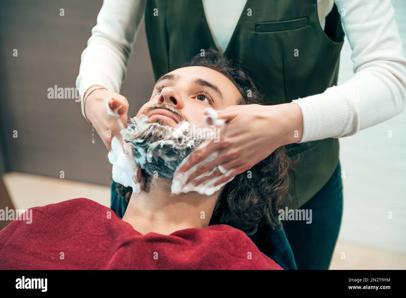 High angle of crop female barber in uniform applying soap on clients beard while preparing for trimming procedure in professional barbershop Stock Photo
