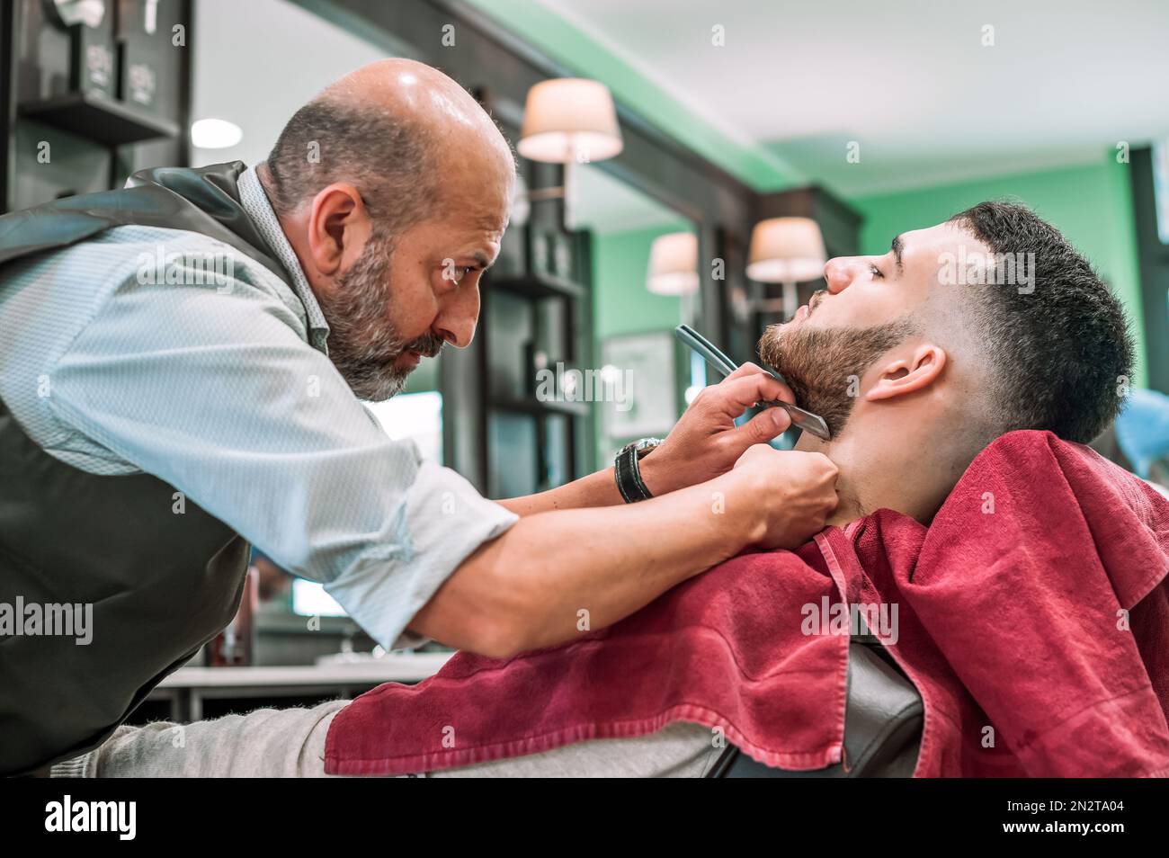 Side view of focused attentive barber using blade shaving razor on male client adjusting beard in barbershop Stock Photo
