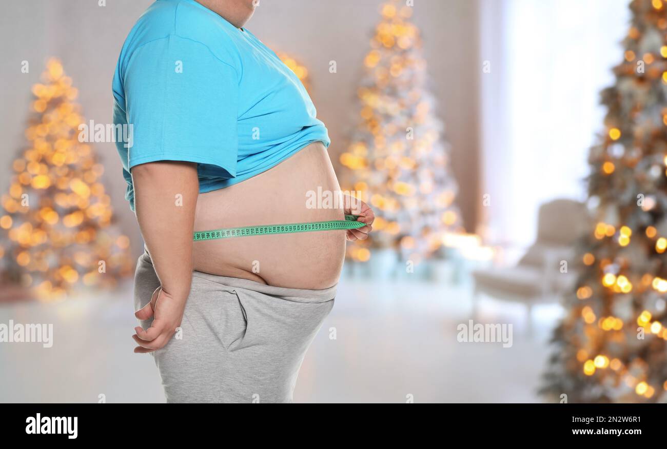 Overweight man measuring his waist in room with Christmas trees after holidays, closeup Stock Photo