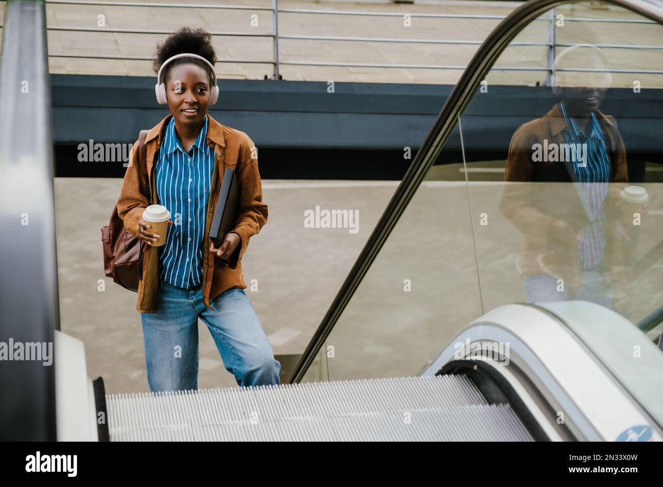 Young beautiful african woman in headphones with coffee and folder looking aside while standing near escalator outdoors Stock Photo