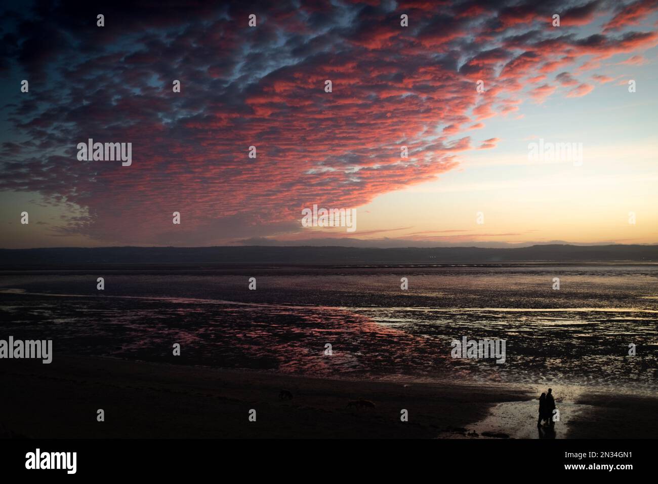 Two people walking along the beach at Cubbins Green, West Kirby, Wirral, looking over to the coast of North Wales on a February evening at sunset. Stock Photo